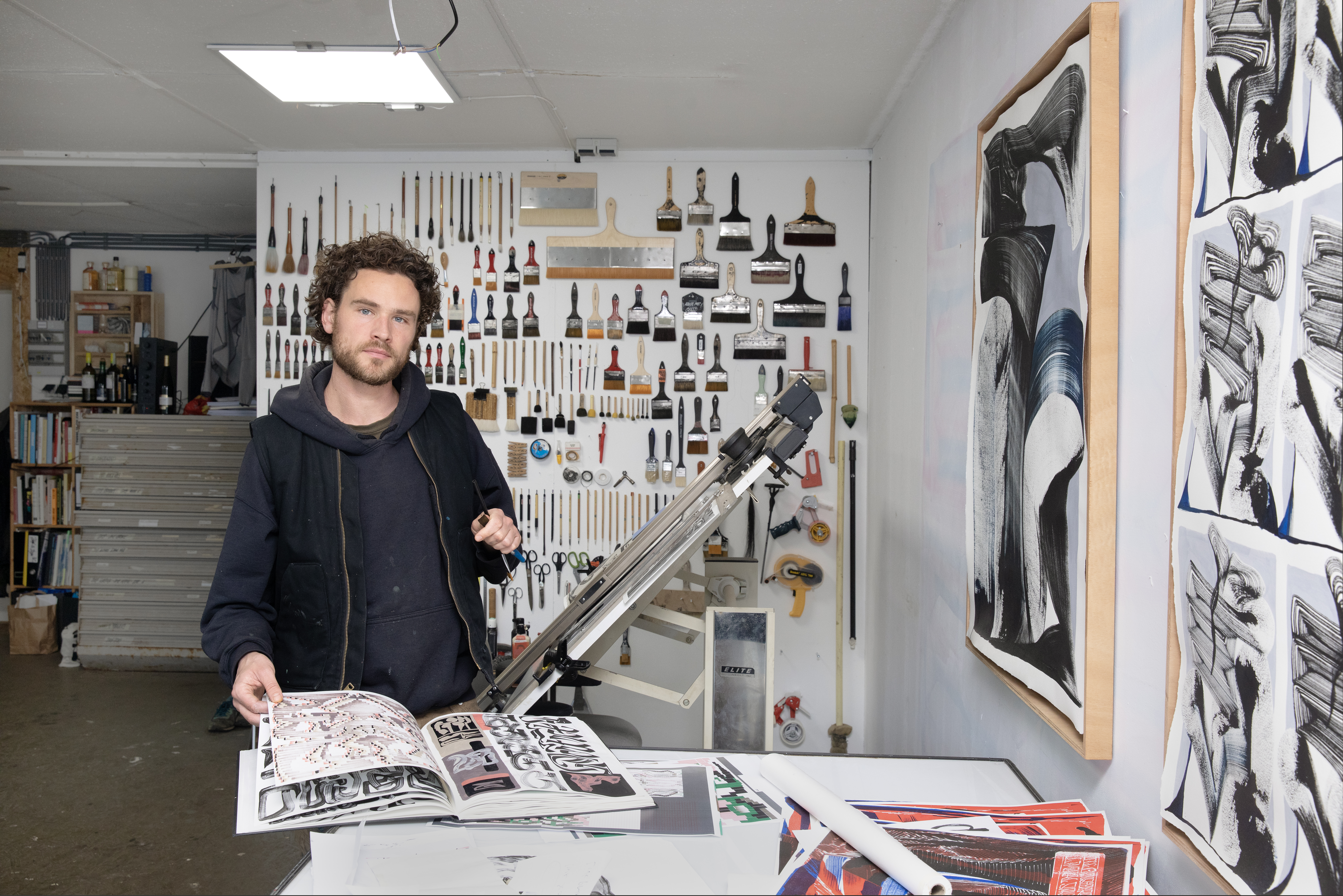 Vincent de Boer staat in zijn atelier aan een werktafel en bladert in een catalogus van zijn werk. Aan de linkerhand hangen werken op papier en recht achter hem een grote collectie penselen aan de muur
