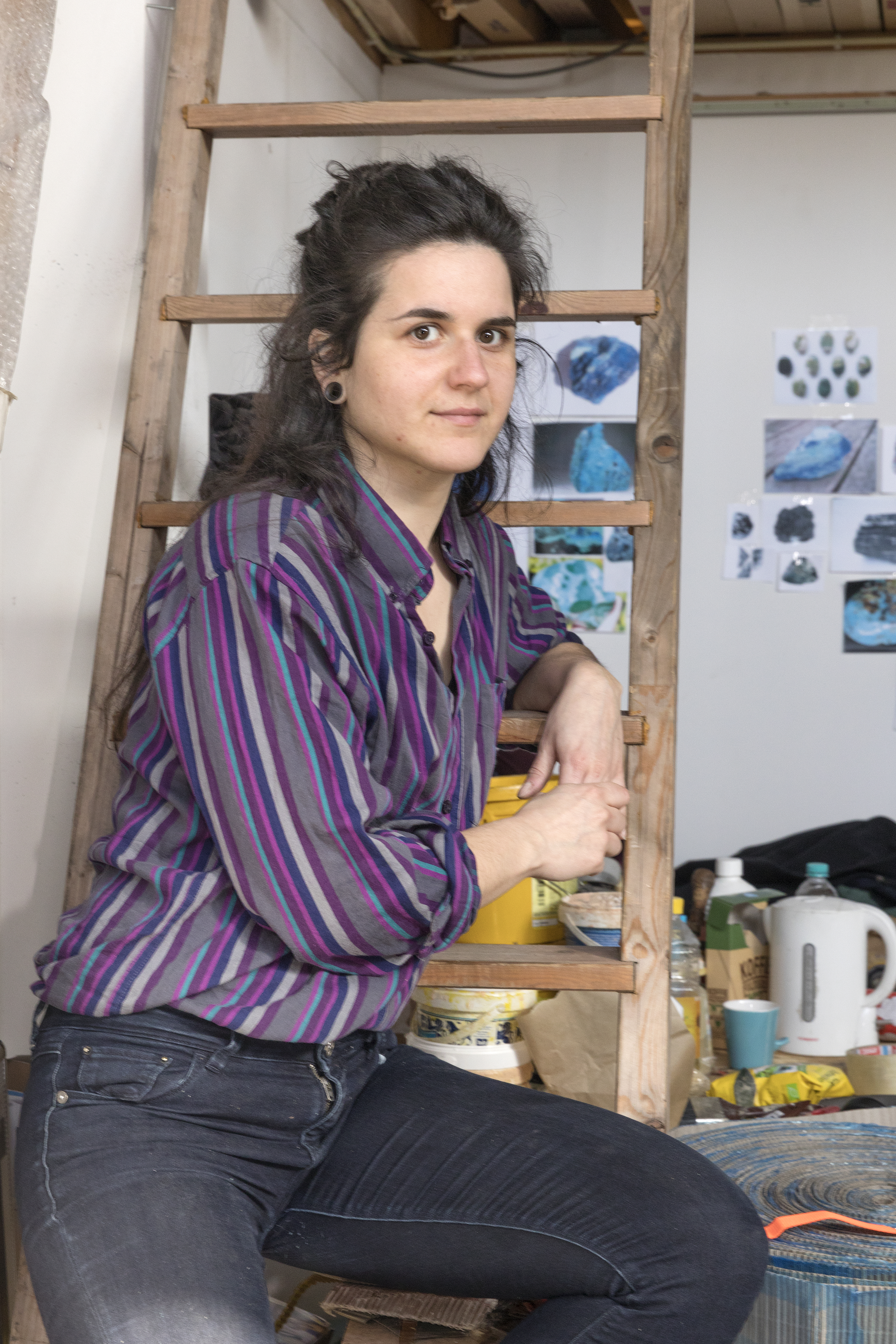 Matea Bakula leunt, gekleed in een paars gestreept overhemd en zwarte spijkerbroek, tegen een ladder in haar atelier. Op de achtergrond zijn materialen en schetsen te zien