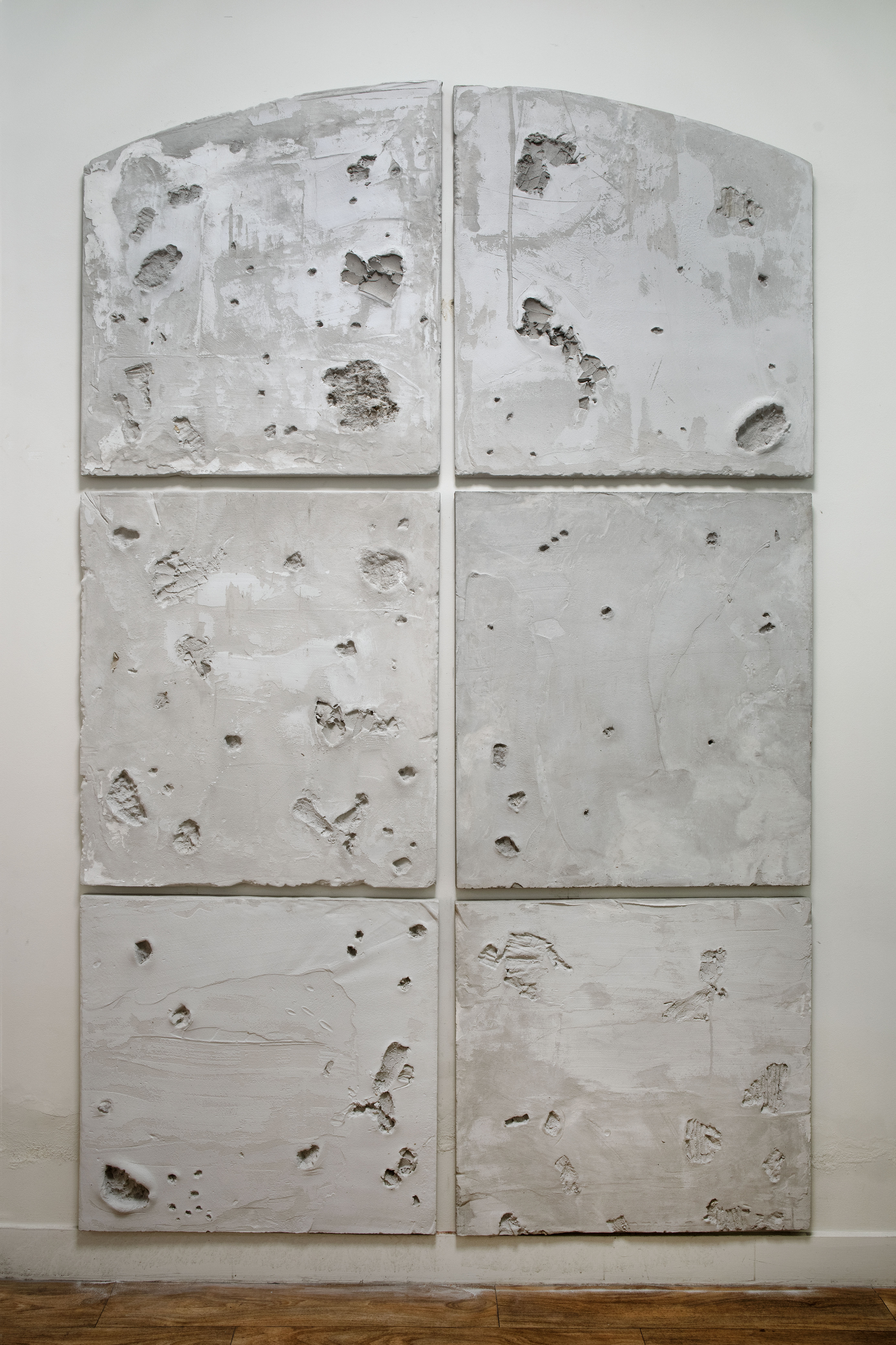 Foto van zes panelen uit het werk ‘dragen’, alsof het een raamvenster is met 6 vensters. De bovenste twee zijn in boogvorm gegoten. De panelen zijn in cement gegoten en bevatten sporen van materialen uit de kelder.