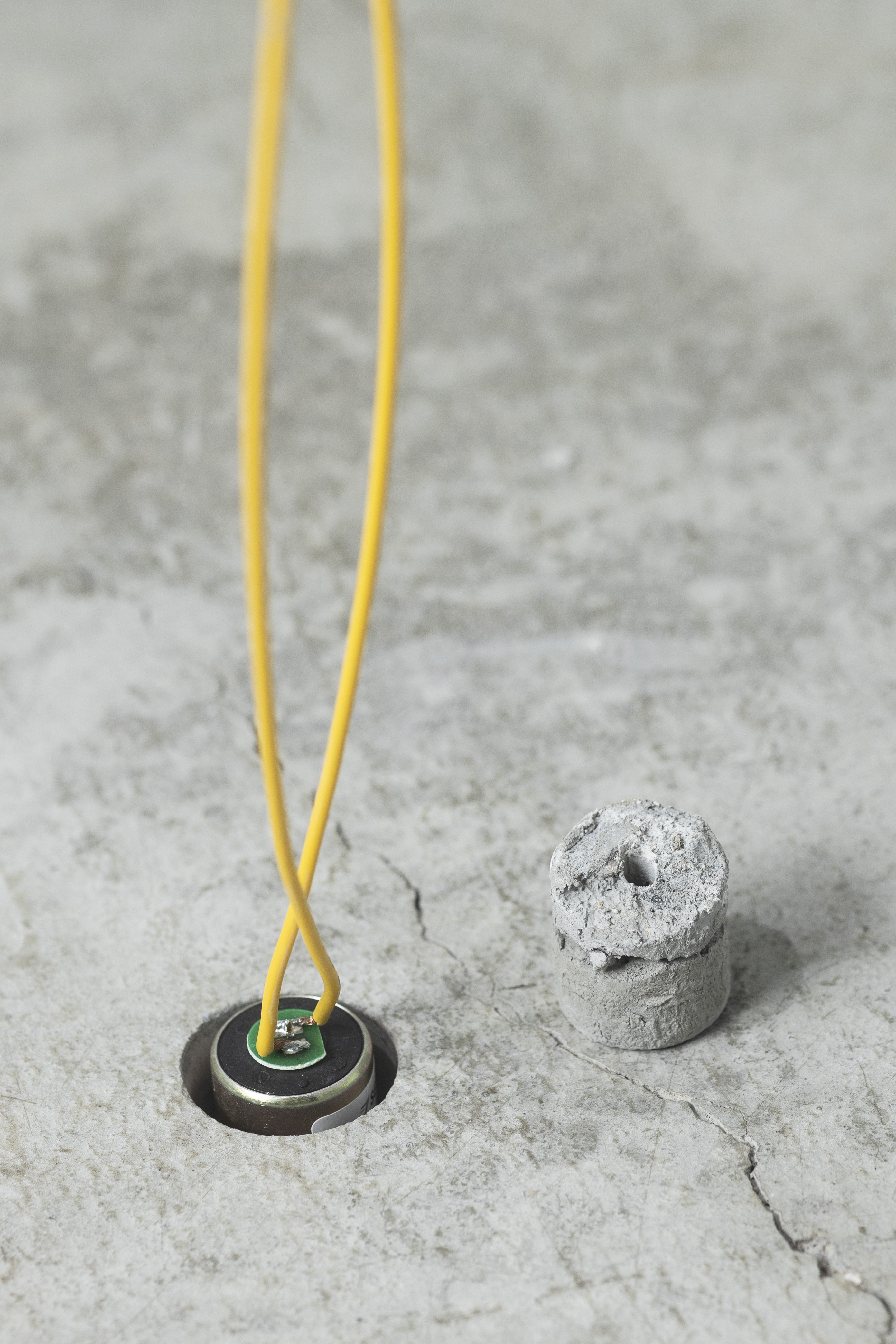 twee gele stroomkabeltjes zijn aangesloten op een batterij die in een gaatje in een stuk beton steekt. Een rond stukje beton (uit het gat) staat los ernaast.