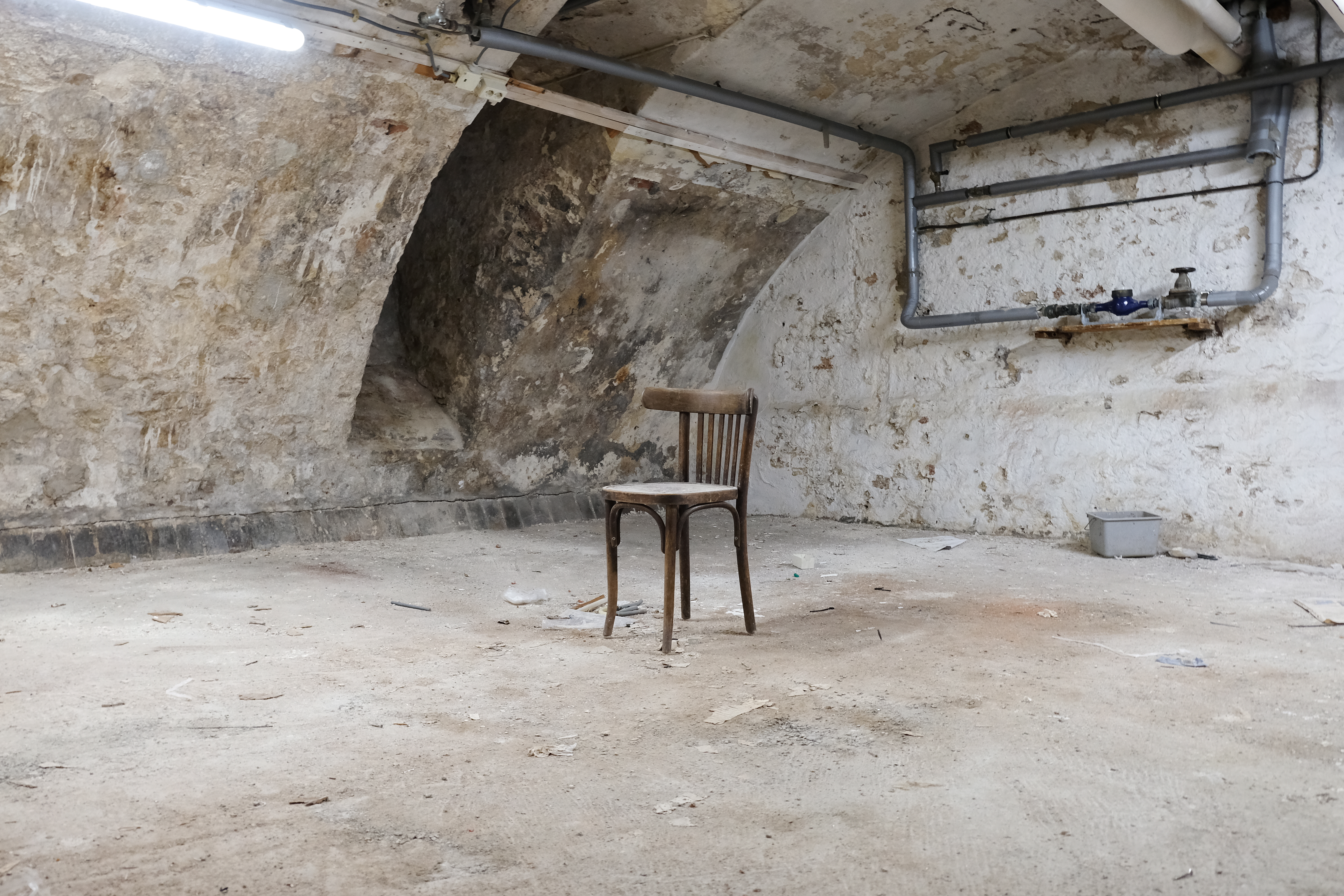 Foto van de kelder. De ruimte is kaal, de vloer en muren bestaan uit stenen en cement. Midden in de ruimte staat een houten stoel onder het stof.