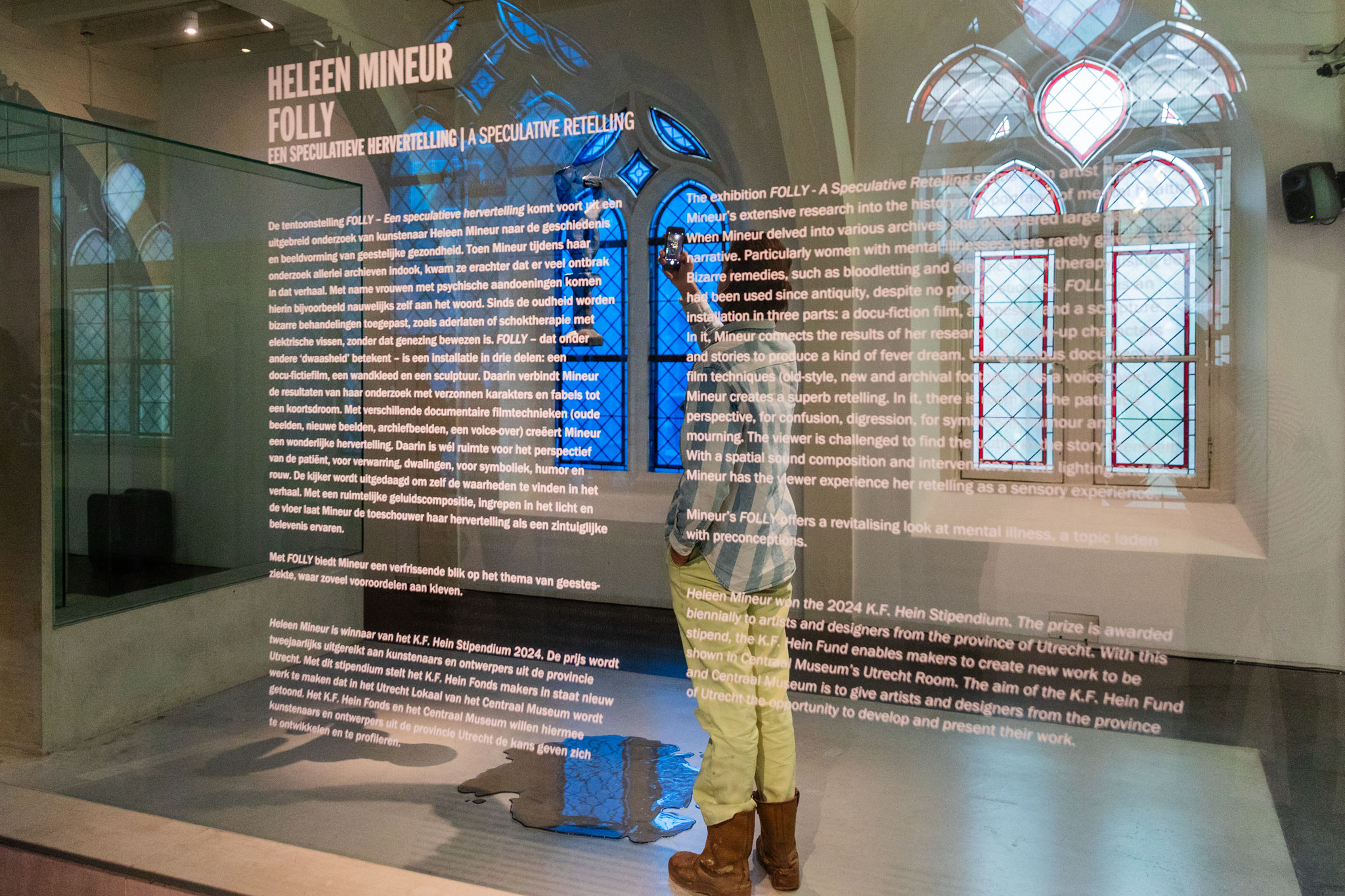 bezoeker fotografeert de sculptuur in de expositie, op de voorgrond de zaaltekst van de tentoonstelling op een glazen wand