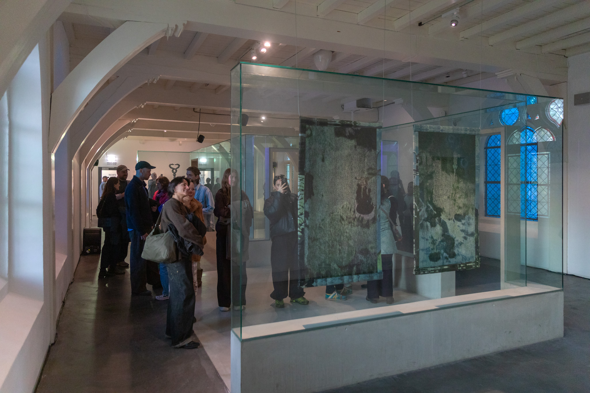 achterzijde van de wandkleden door de glazen tentoonstellingswand heen gezien, publiek bekijkt en fotografeert de kleden