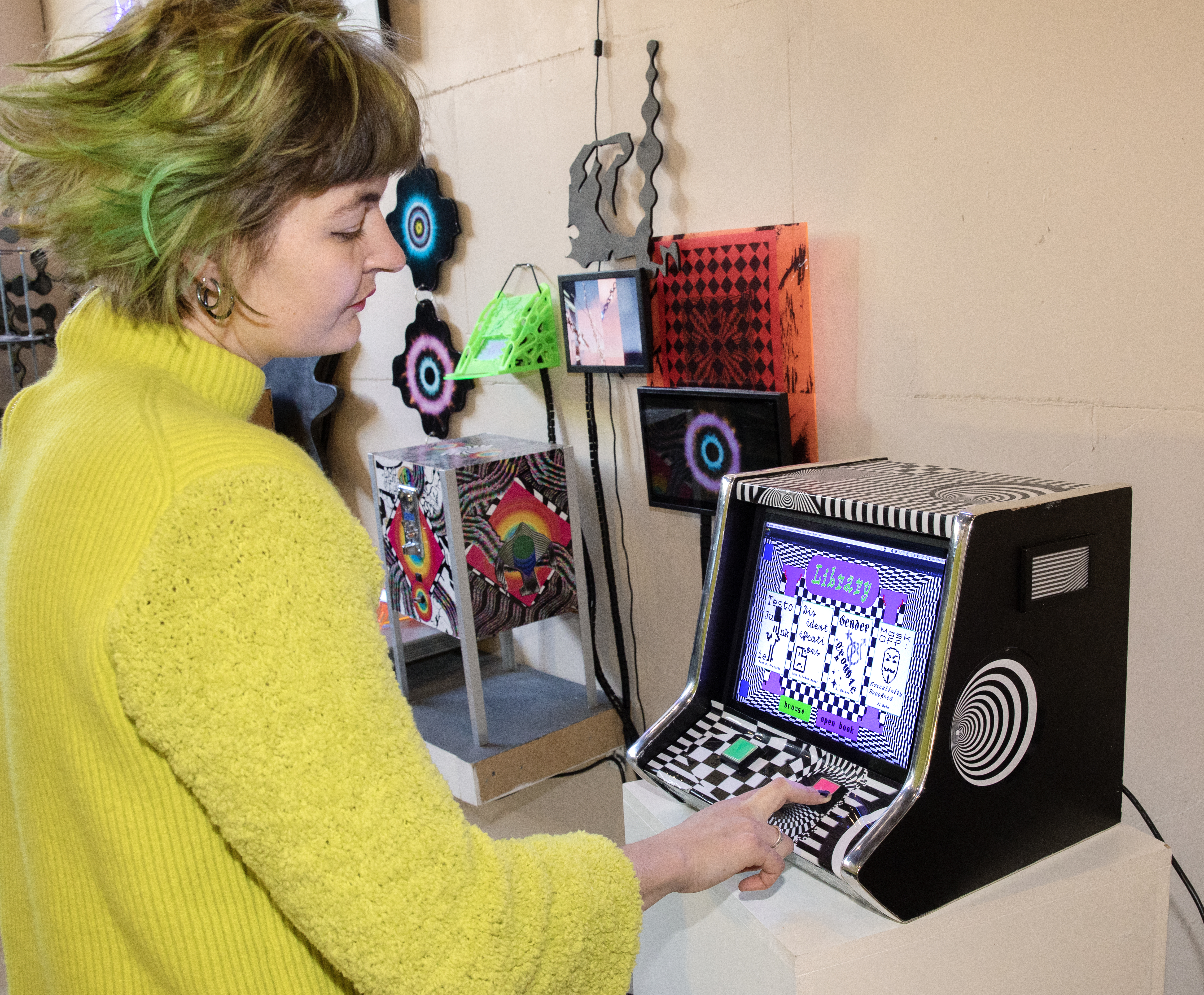 Kunstenaar Cyanne van den Houten (gele trui, groen kort haar) demonstreert haar prototype game op een ouderwetse spelcomputer met twee knoppen. De spelcomputer is vormgegeven als het spel, met een grafische zwart-wit print.