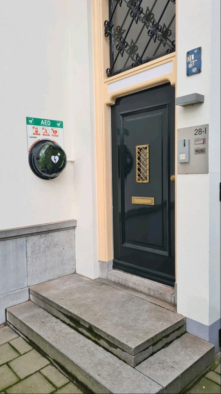 De AED hangt links aan de buitenmuur naast de ingang van het kantoorpand bij nummer 28-1. Op de kast van de AED staat een hart met een bliksemschicht en een plusje. Daarboven hangt een bord met drie pictogrammen hoe de AED gebruikt moet worden